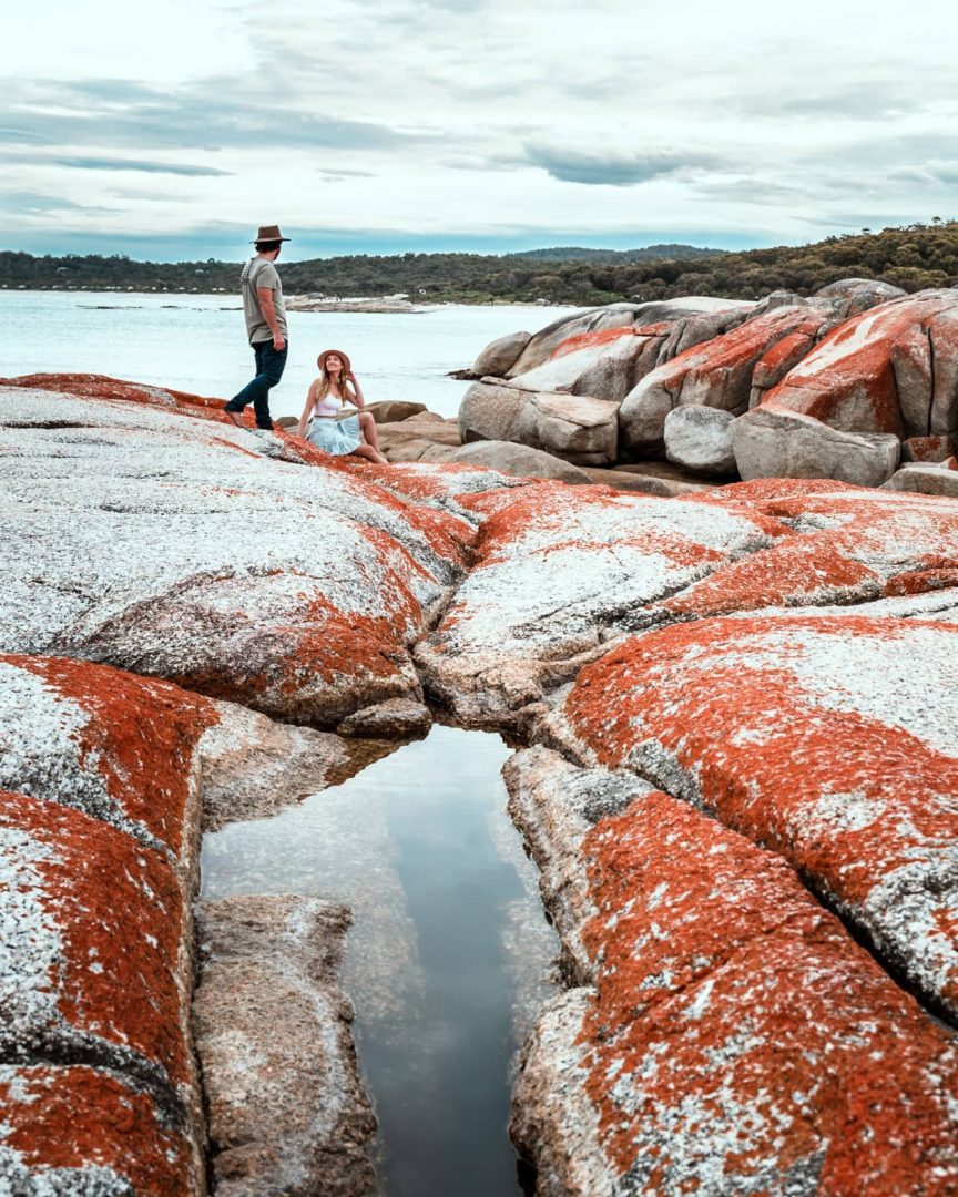 A couple explores vibrant orange and white lichen-covered rocks at the Bay of Fires, a popular location on a 7 day Tasmania road trip.