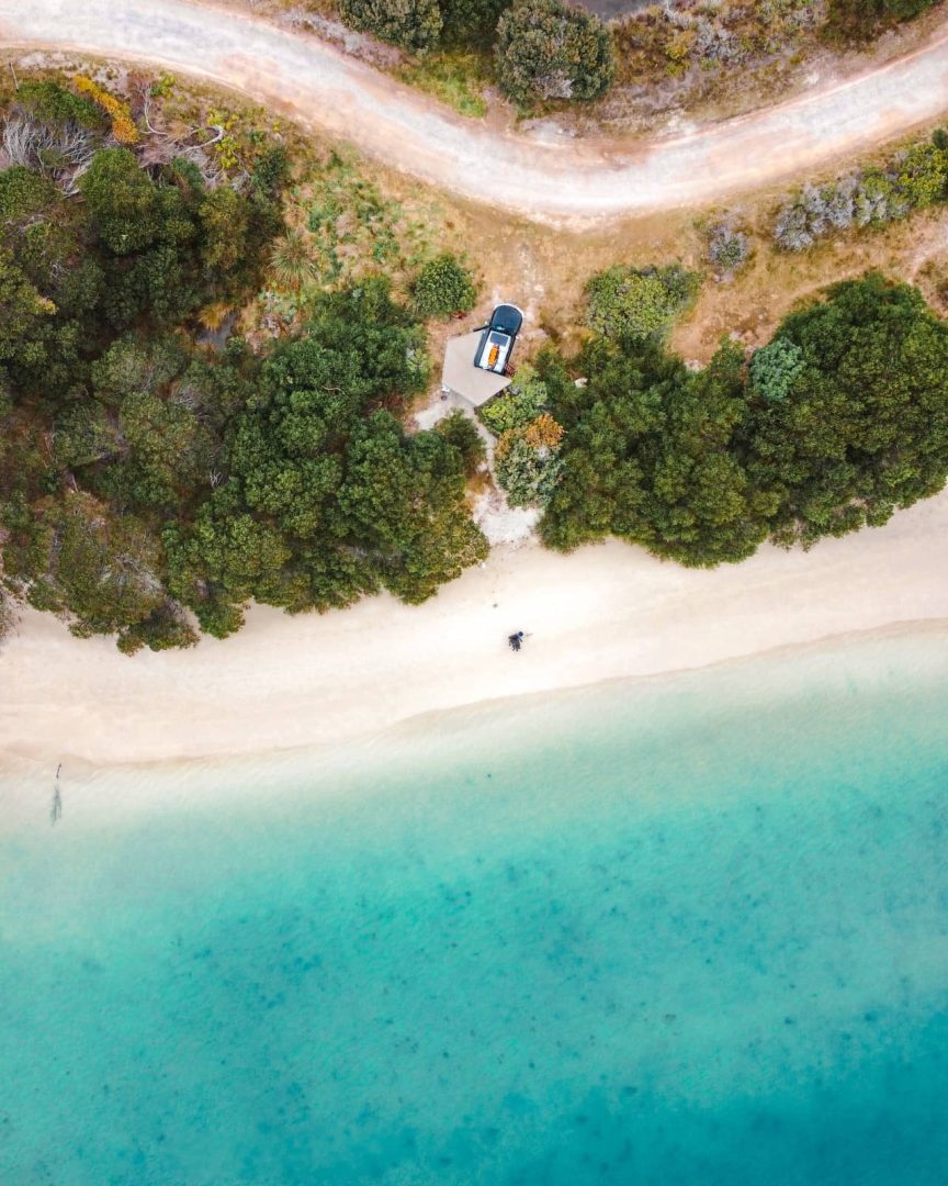 Aerial view of a camper parked near dense bushland beside a pristine white sand beach with turquoise water, capturing a serene moment on a Tasmania Road Trip 10 Days.