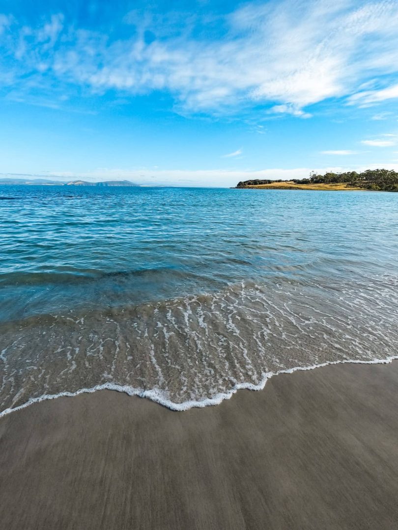 Small wave washing over smooth sand under a blue sky at a peaceful cove along the Tasmania East Coast Road Trip.