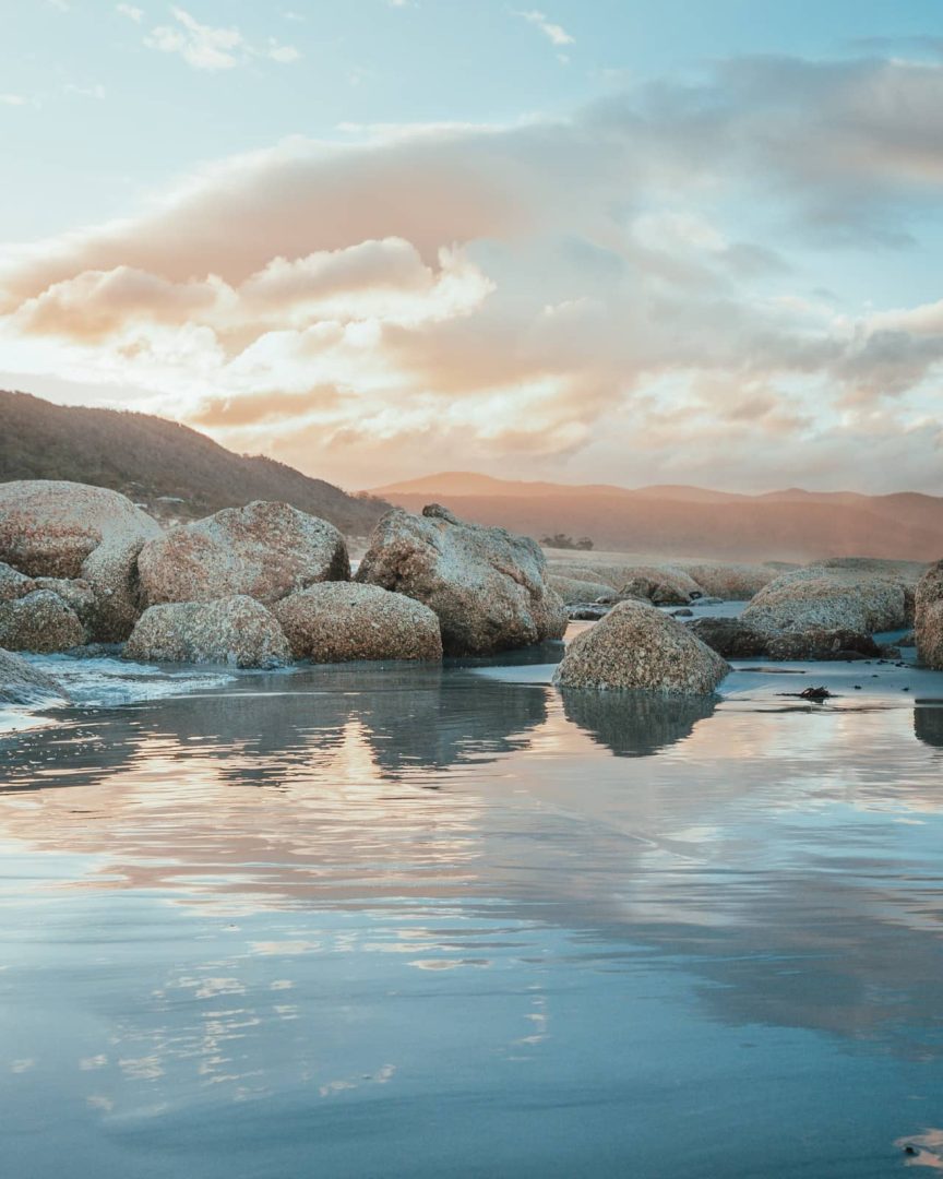 Granite boulders on Diamond Island, near Bicheno, reflecting in the wet sand at sunset with soft orange light and misty hills in the distance, captured on the great eastern drive.