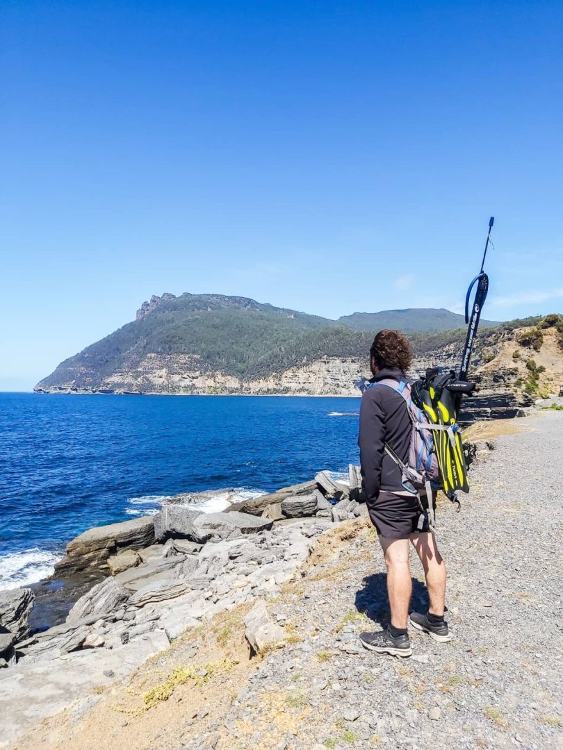 Man with a spearfishing gun strapped to his pack standing on a rocky coast, looking out over the deep blue sea during a coastal stop on a Tasmania Road Trip 10 Days.
