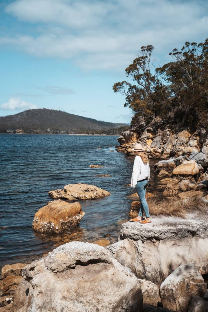 Woman standing on coastal rocks looking out over calm water and distant hills, enjoying a quiet moment on one of the scenic things to do in Tasmania.