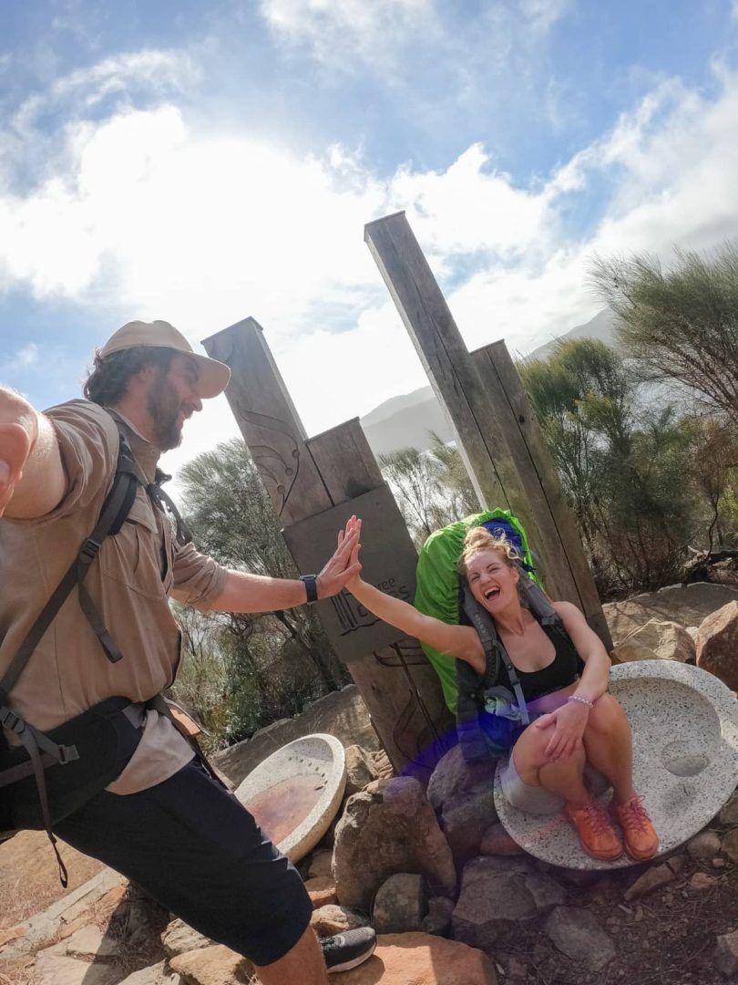 Two hikers celebrate with a high-five at the Three Capes Track finish, next to the tall timber pillars marking the end of their free hike.