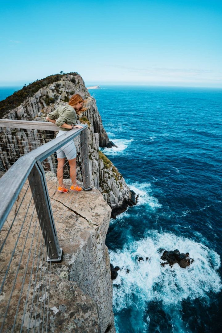 A woman leans over a clifftop lookout railing above wild ocean swells and jagged rocks, on Cape Hauy, a thrilling highlight on the Three Capes Track for free.