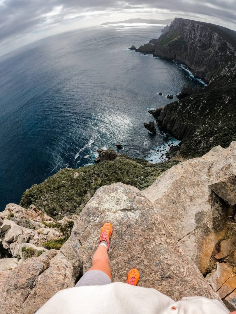 A hikerโs feet in orange shoes stand at the edge of a rocky cliff high above the sea, capturing the vertigo-inducing heights on the Three Capes Track for free.