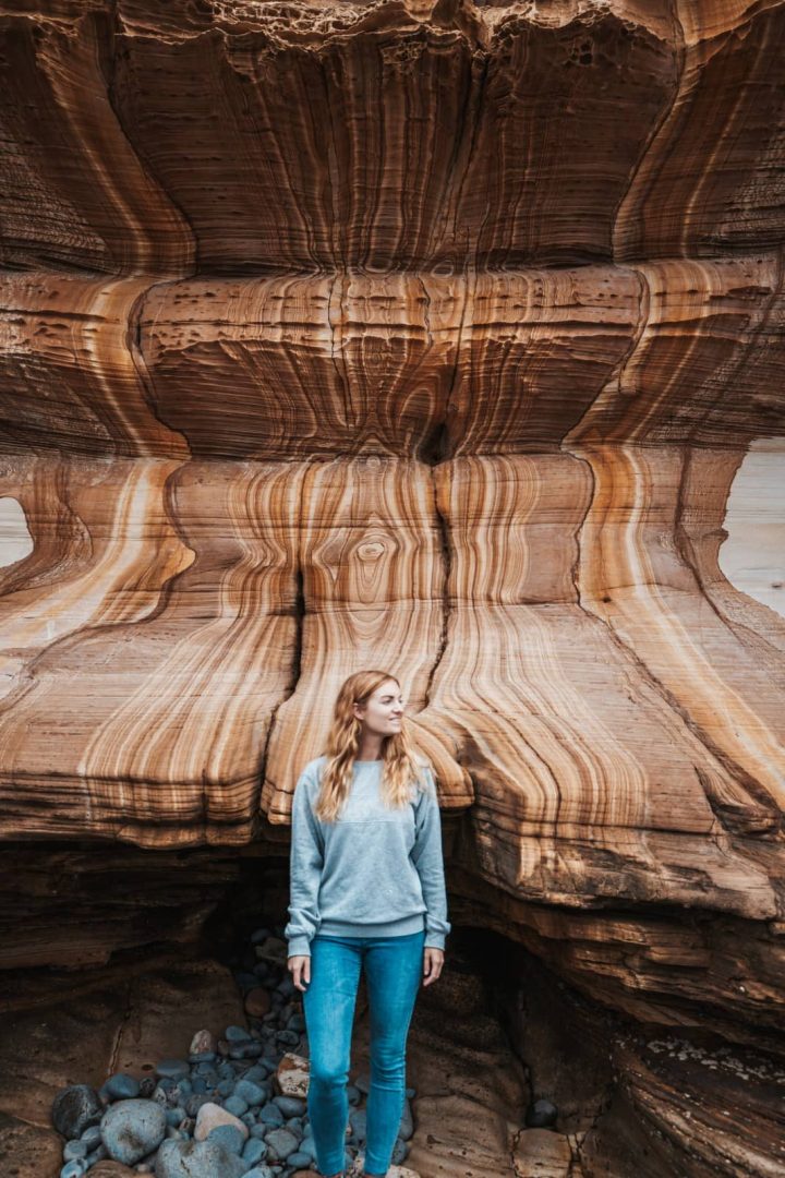 Woman standing in front of the Painted Cliffs, a striated orange and brown patterned rock wall on Maria Island, a geological wonder and one of the unique things to do in Tasmania.