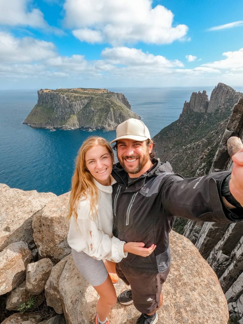 A couple smiles at the camera with Tasman Island and dramatic cliffs behind them, a highlight of hiking the Three Capes Track for free.