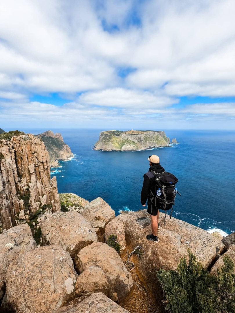 Solo hiker with a backpack looking out over Tasman Island from a cliffโs edge on a clear day, after doing the Three Capes Track, capturing the adventurous side of things to do in Tasmania.