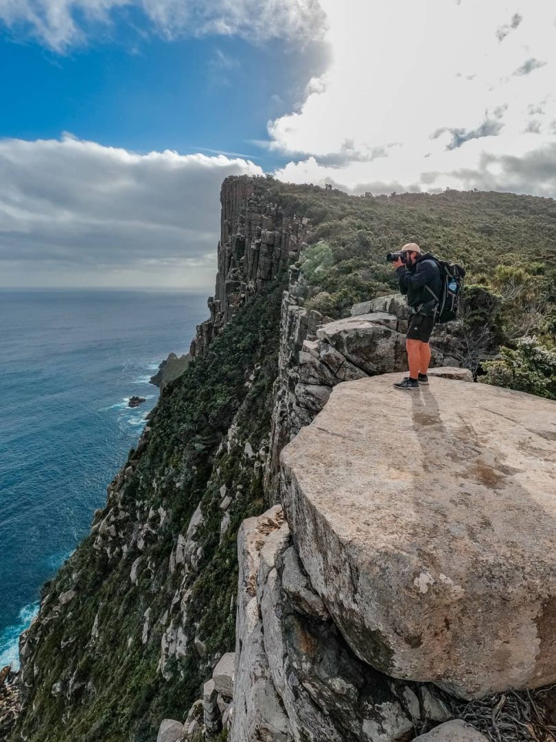 A photographer stands on a rocky ledge aiming a camera at the coastline, documenting the dramatic landscapes of the Three Capes Track for free.