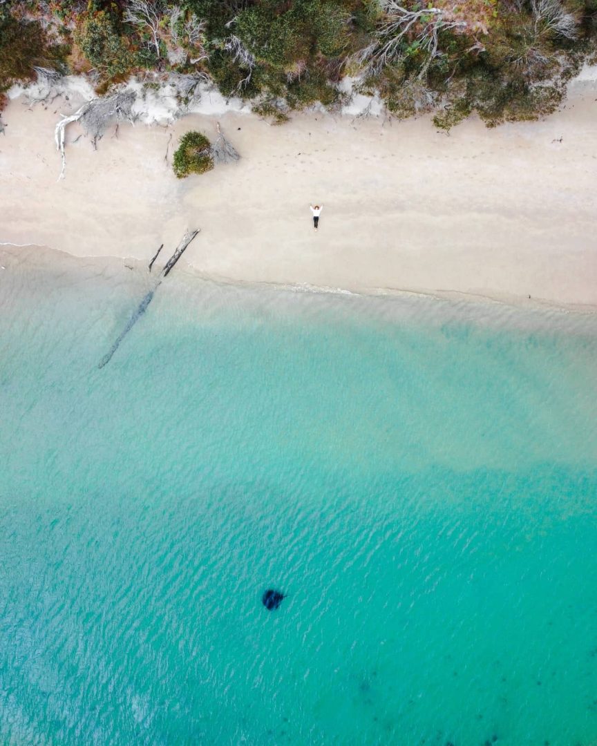 Aerial view of a person lying on a quiet, sandy beach on Cockle Creek,beside clear turquoise waters and forest edge, capturing the peaceful coastal side of things to do in Tasmania.