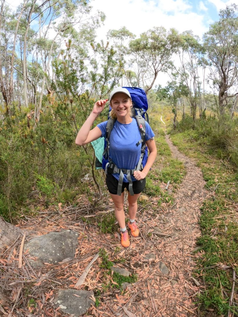 A female hiker smiles on a narrow bush track lined with gum trees, trekking through the forested section of the Three Capes Track for free.
