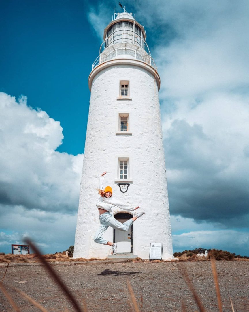 Person mid-jump in front of Cape Bruny lighthouse under a dramatic sky, one of the quirky and photogenic things to do in Tasmaniaโs coastal areas.