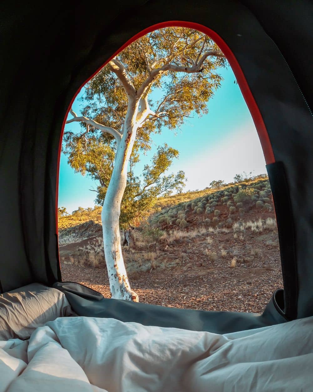 View from inside a tent looking past white bedding toward a pale gum tree and rocky red landscape in Karijini National Park. It shows what waking up inside the park can look like when staying near the gorges.