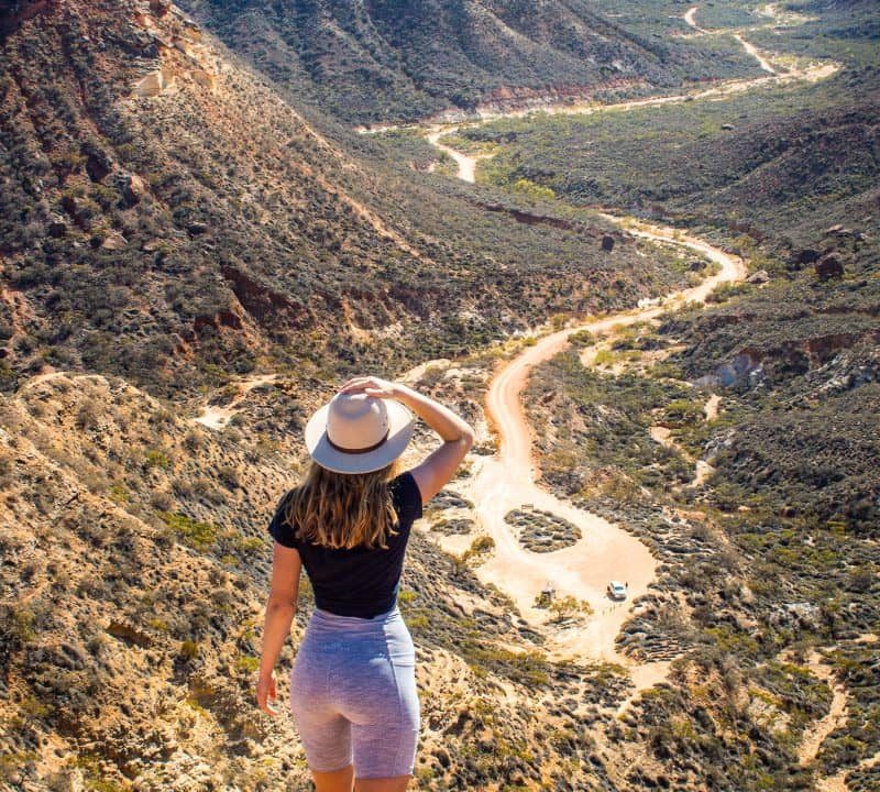 Beck of Salt and Charcoal gazes over Shothole Canyon in Western Australia, standing on a rugged overlook with her hand shielding her eyes from the sun, surveying the winding dirt road that carves through the arid valley below.