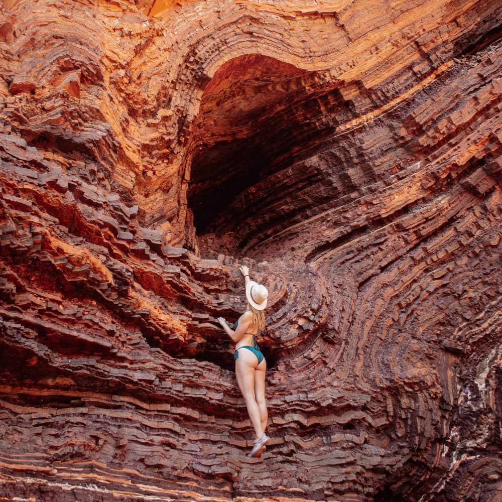 A woman in swimwear and a hat stands against the richly layered red rock formations in Pilbara, extending an arm to touch the ancient, curved sedimentary walls.