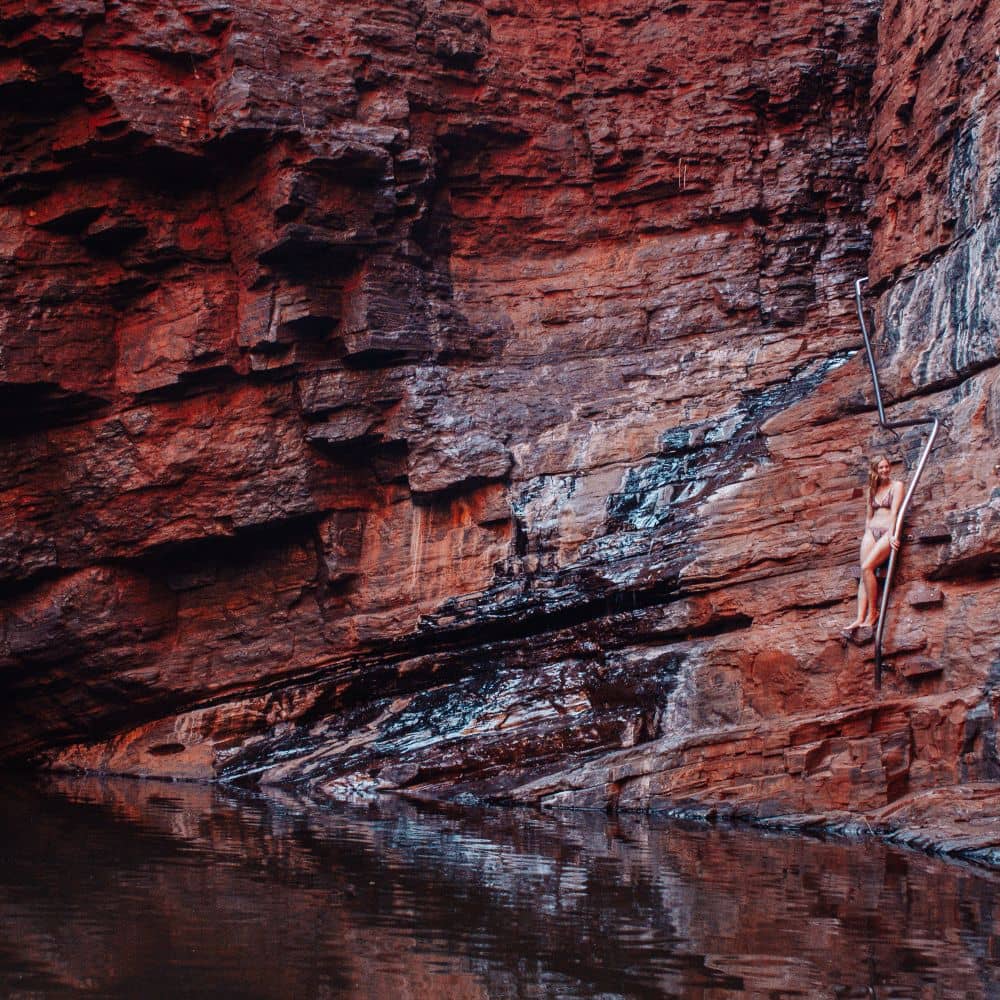 Woman standing on rock, holding rail while smiling and looking at the body of water beneath her. Located at the Handrail Gorge in Karijini. 