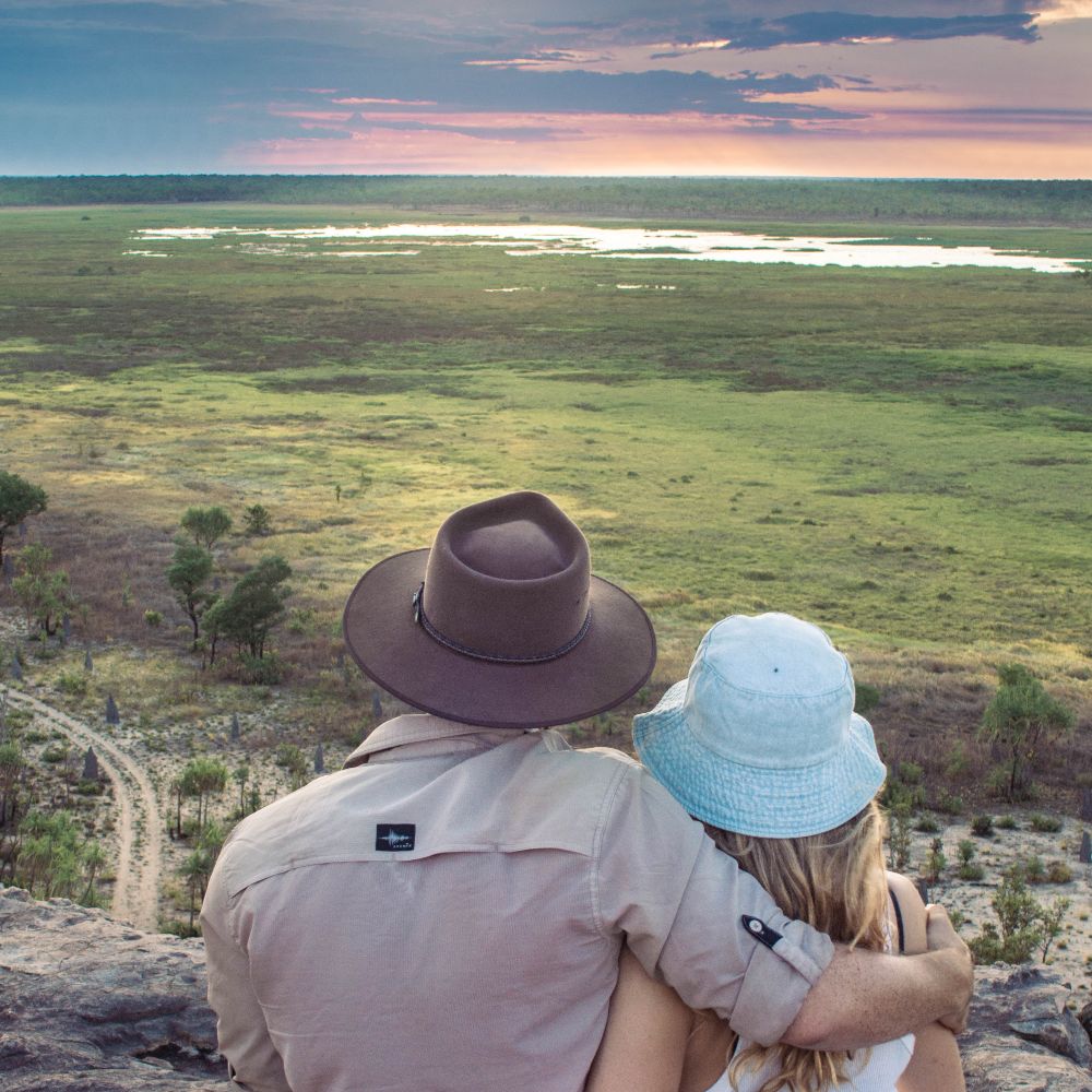 A couple sits close together on a rocky Ubirr lookout as they watch the wetlands beneath a pastel evening sky. The broad floodplain view is a memorable stop on a Darwin to Kakadu weekend road trip.