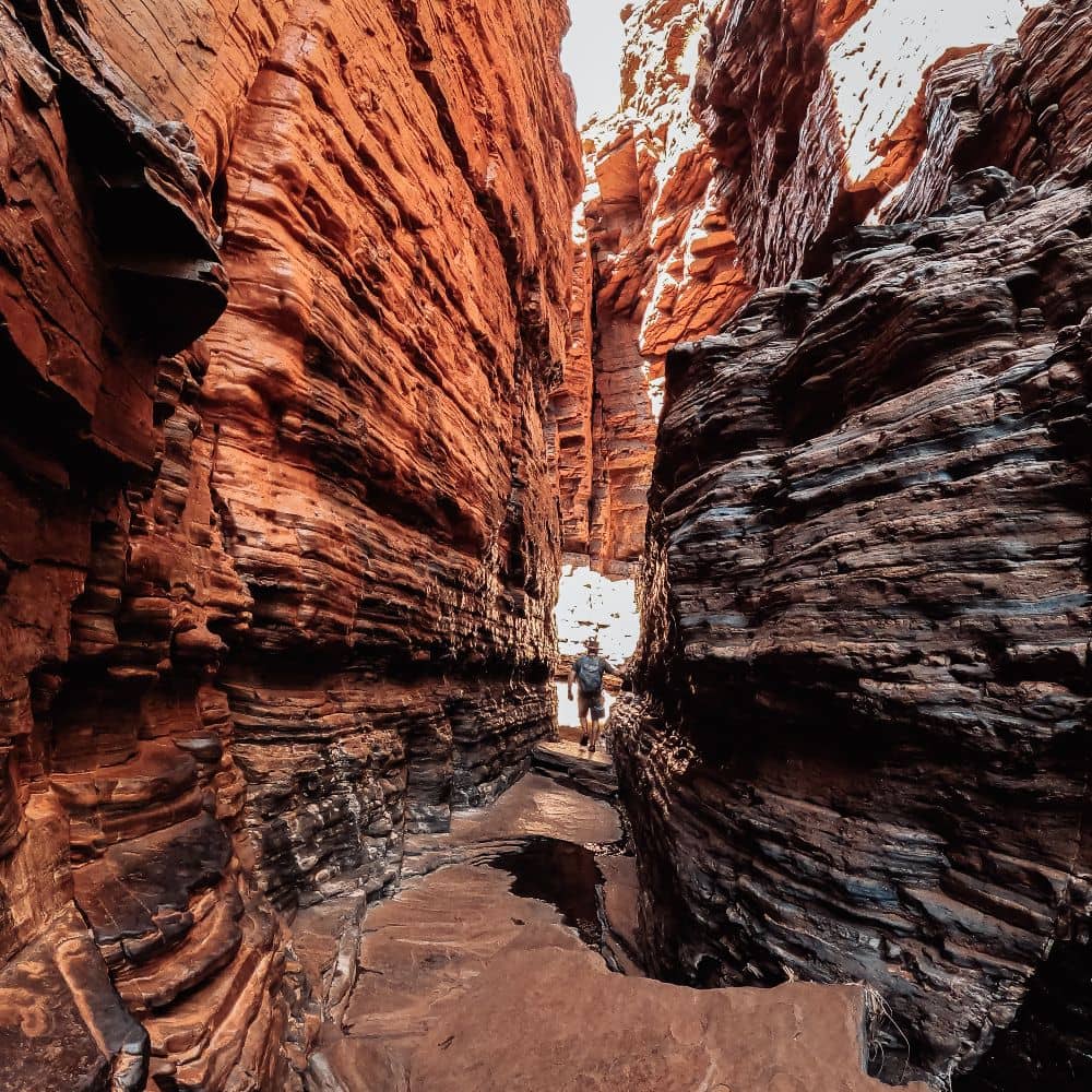 Man walking through a gorge surrounded by red rocks. One way of walking through the Weano Gorge in Karijini, specifically the Handrail pool descent and discovering it's rich history.