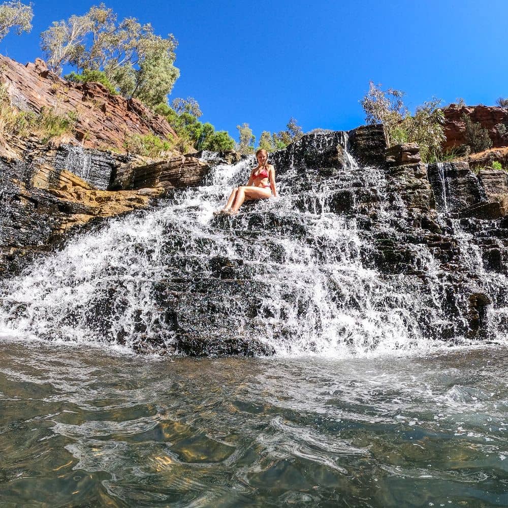 Women sitting on waterfall as water gushes down to the body of water below. One of the best things to do in Karijini. Can be found at the Fortescue Falls, Karijini. 