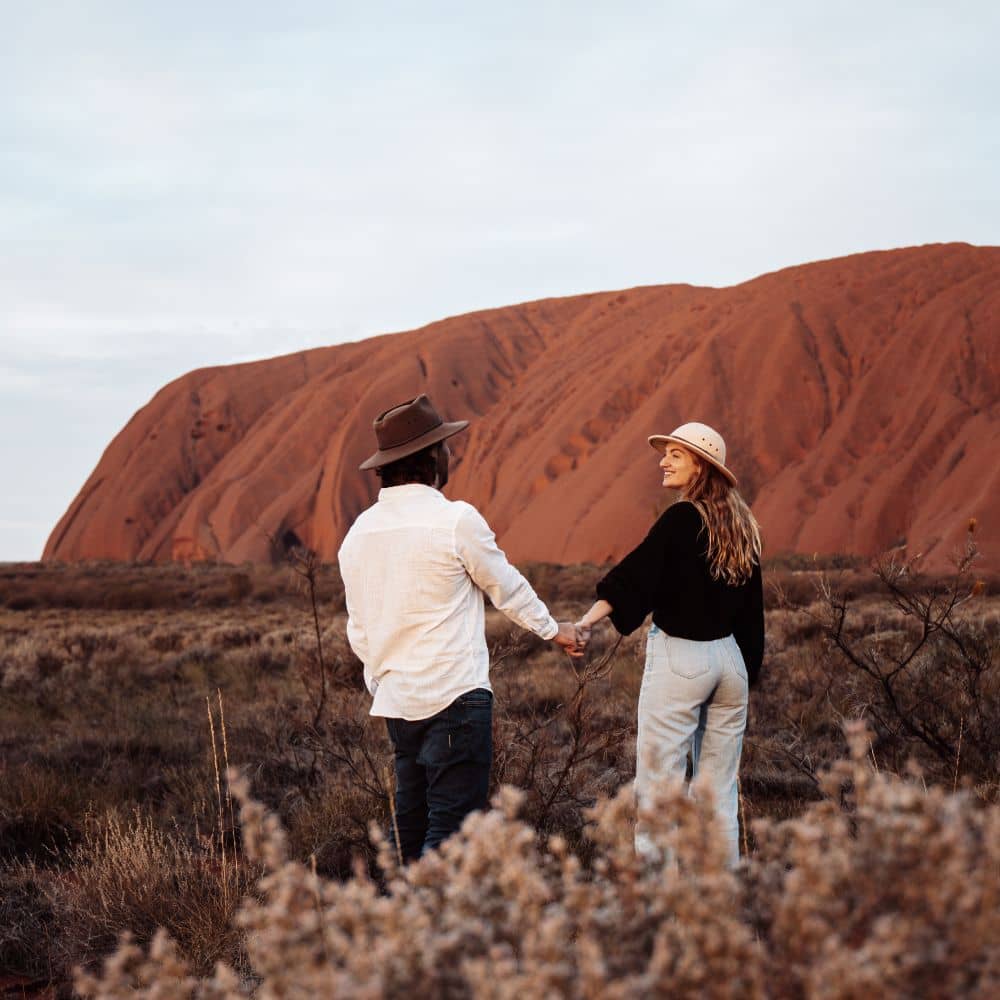 Man and woman holding hands while looking at the Uluru. An insiders guide to when to go to Uluru for the best experience. This is at the red centre of Australia, Uluru located in the Northern Territory. 