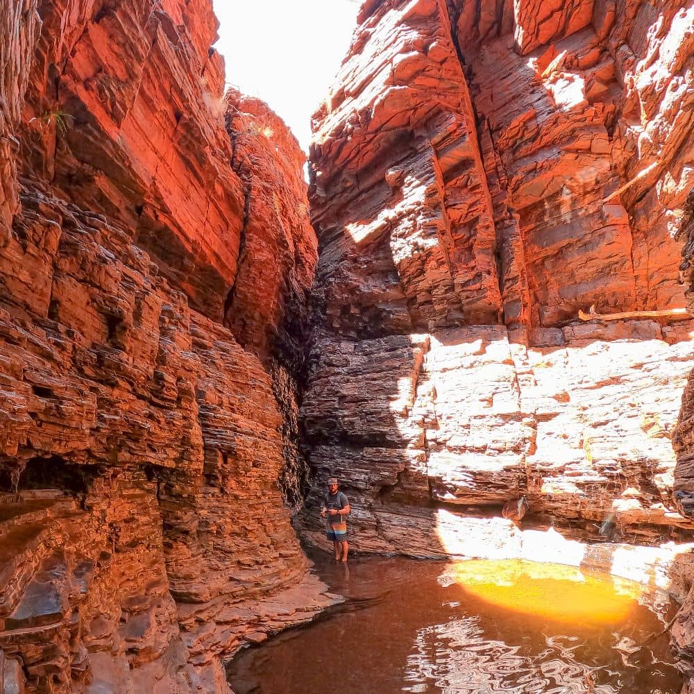 Man standing in gorge surrounded by red rocks with feet half in water. Can be found in the gorgeous Weano Gorge in Karijini, at the Handrail pool hike.