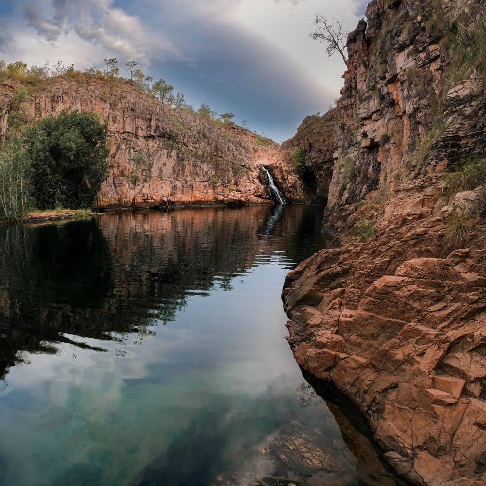 A river with a waterfall and rainbow overtop. This is just one of 13 unmissable activities to see in Kakadu National Park. This is the Barramundi Gorge/Majuk Falls. 