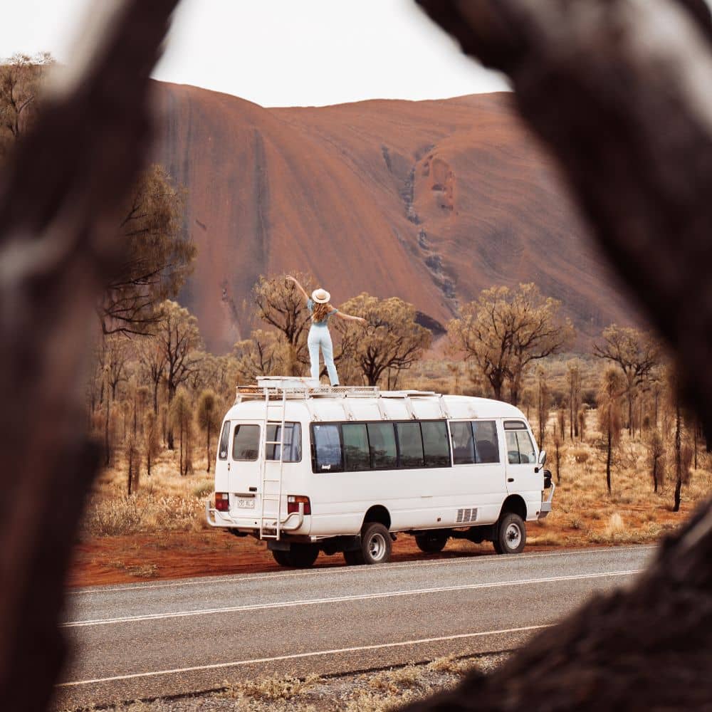 Woman standing on top of a van while looking at Uluru. You need the ultimate guide to know when to go to Uluru. This is at Uluru, NT.