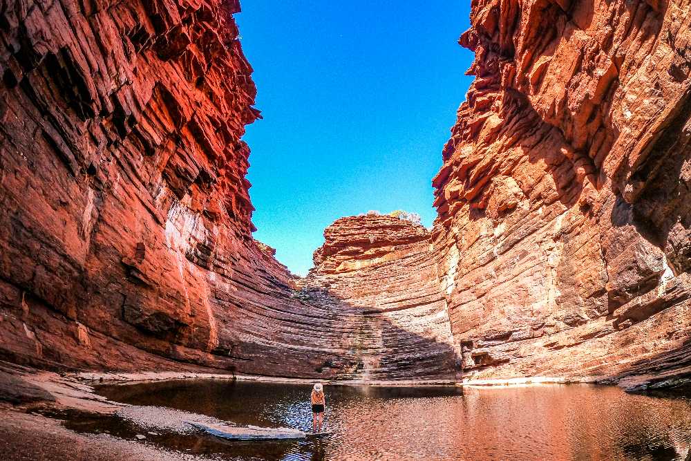 Beck of Salt and Charcoal walk through the expansive Hancock Gorge within Karijini National Park, Western Australia. The gorge walls rise dramatically in layers of deep red and orange rock, encasing a still pool of water reflecting the vibrant colors and the brilliant blue sky above.