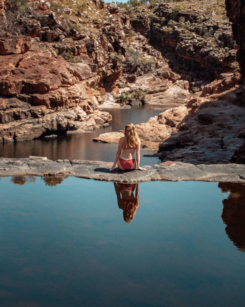 A woman sits on a flat rock overlooking a peaceful, reflective pool at the top of Bell Gorge, with red cliffs and rugged terrain stretching into the distance—one of the most photogenic spots along a gibb river road itinerary.