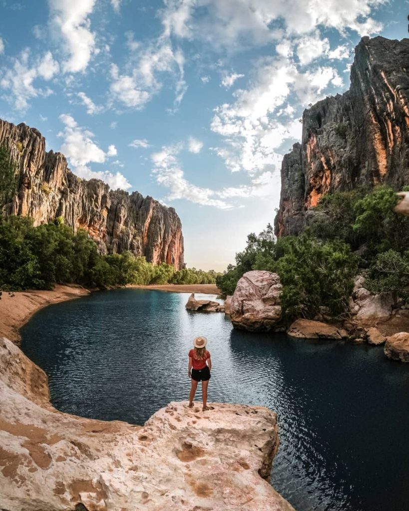 A woman stands on a rock ledge overlooking the still waters of Windjana Gorge, surrounded by towering limestone cliffs and lush vegetation—an unmissable natural wonder on a gibb river road itinerary.