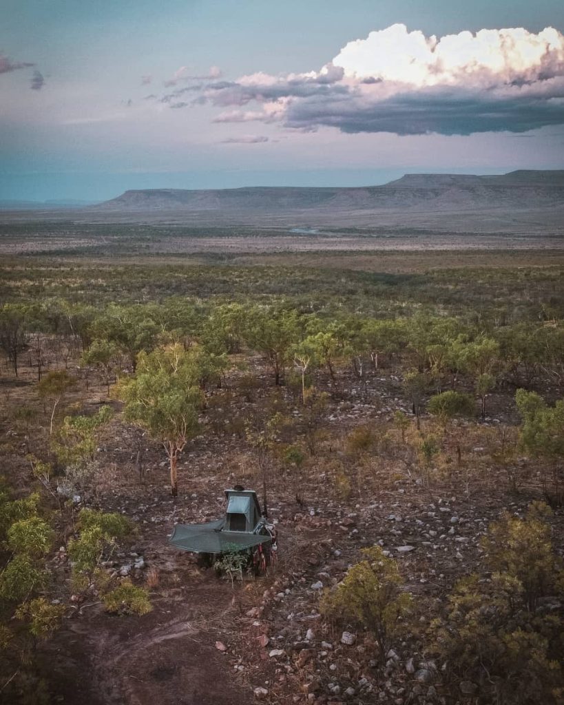 A 4WD with a rooftop tent set up in a rocky clearing surrounded by sparse bushland and hills at sunset—typical of free camping options along a gibb river road itinerary.