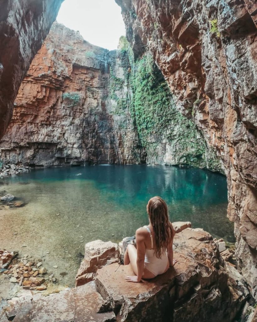 A woman in a white swimsuit sits on a rocky ledge overlooking the turquoise plunge pool beneath a tall, vine-draped cliff at Emma Gorge—one of the most scenic stops along a gibb river road itinerary in El Questro, WA.
