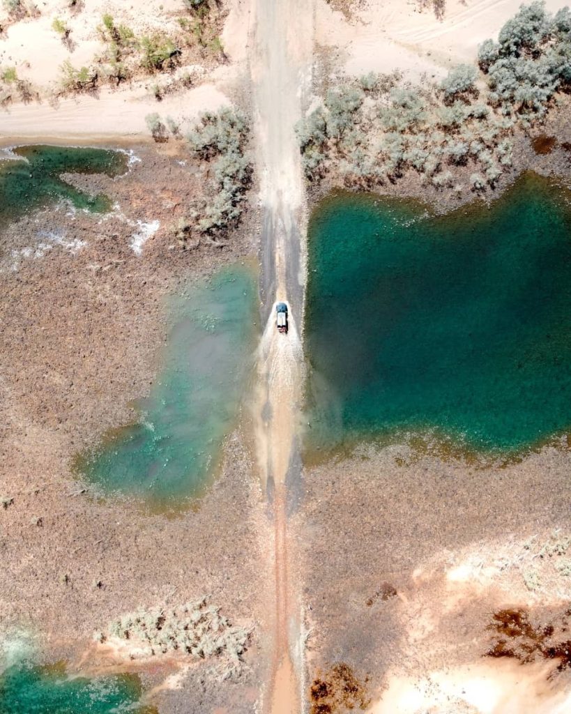Aerial view of a 4WD crossing the Pentecost River, water spraying out on either side, with vibrant turquoise pools and arid terrain—one of the most thrilling highlights on a gibb river road itinerary.