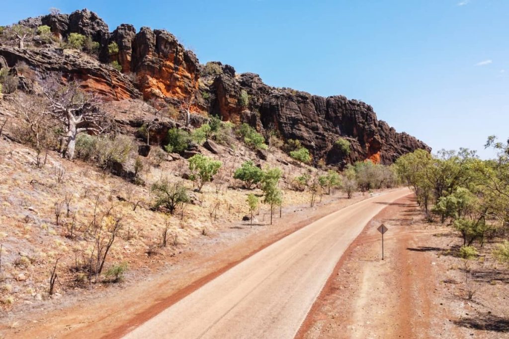 A winding section of the Gibb River Road passes through rugged terrain with red rock cliffs, sparse bushland and boab trees under a clear blue sky—typical scenery featured in a gibb river road itinerary.