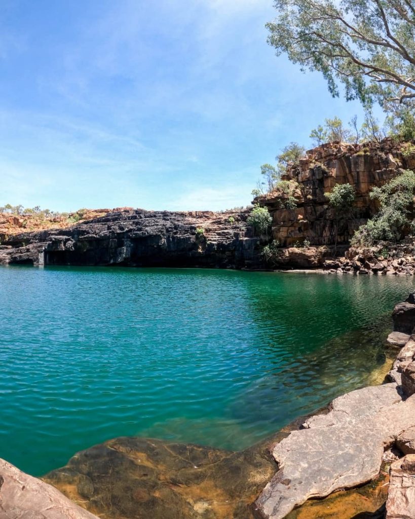 View of the emerald waterhole at Manning Gorge, surrounded by rocky cliffs and sparse vegetation, with a clear sky overhead—a must-see destination on any gibb river road itinerary in the Kimberley.