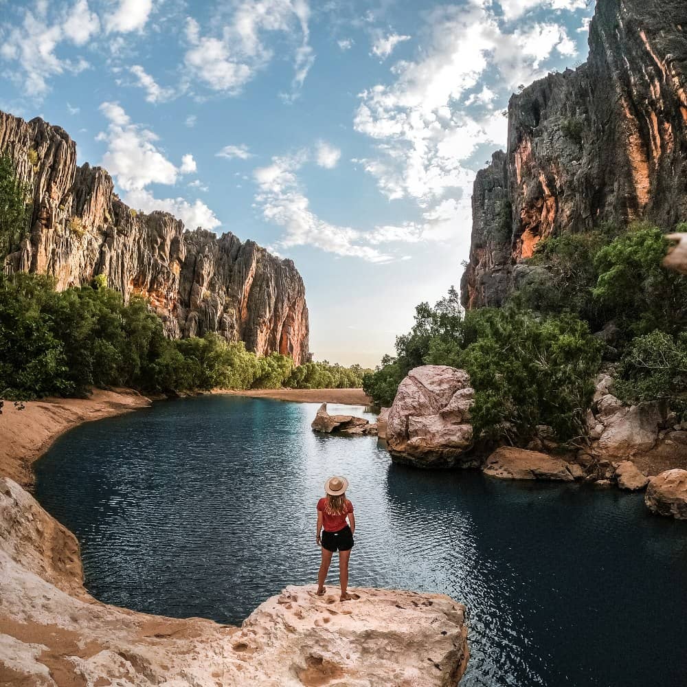 A traveler stands on the edge of a rocky outcrop at Windjana Gorge, Gibb River Road, Western Australia, overlooking a tranquil river flanked by towering cliff walls and lush vegetation.