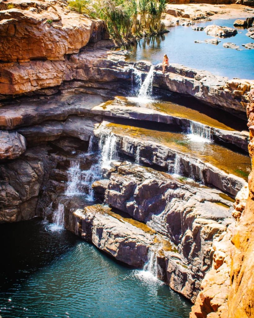 A woman sits on a rocky ledge above cascading tiered waterfalls at Bell Gorge, surrounded by red cliffs and clear plunge pools—an iconic stop on any Gibb River Road itinerary through the Kimberley region.