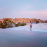 A woman stands in an infinity pool overlooking Lake Argyle near Kununurra. The calm water blends into the lake with rocky hills glowing pink in the distance.