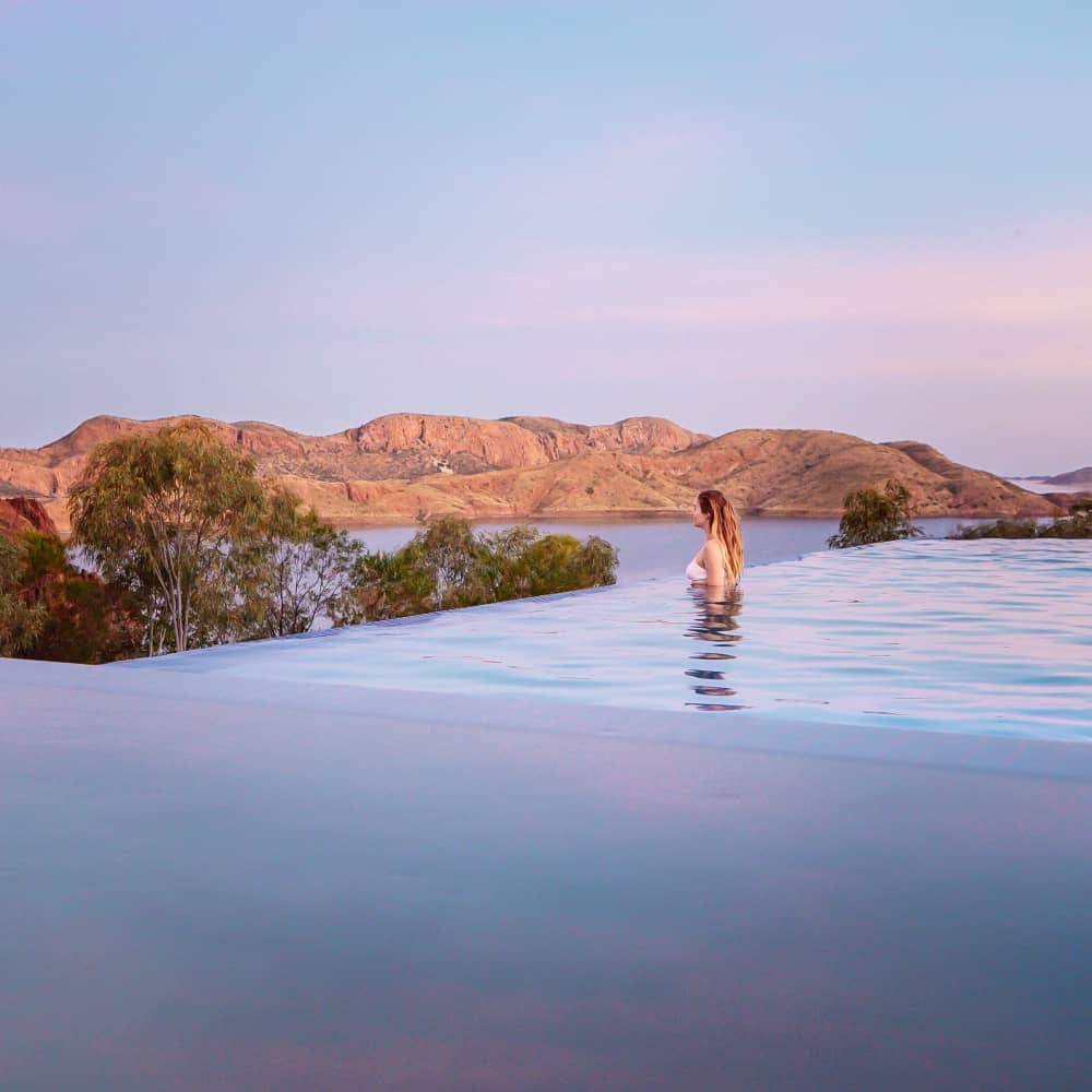 A woman stands in an infinity pool overlooking Lake Argyle near Kununurra. The calm water blends into the lake with rocky hills glowing pink in the distance.