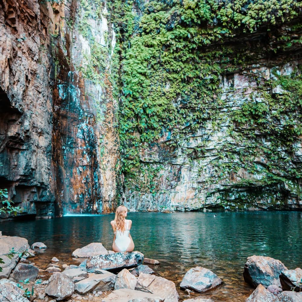 A woman in a white swimsuit sits on a rock at the edge of the swimming hole at Emma Gorge. A waterfall drops into the deep green pool beneath towering cliffs covered in lush plants