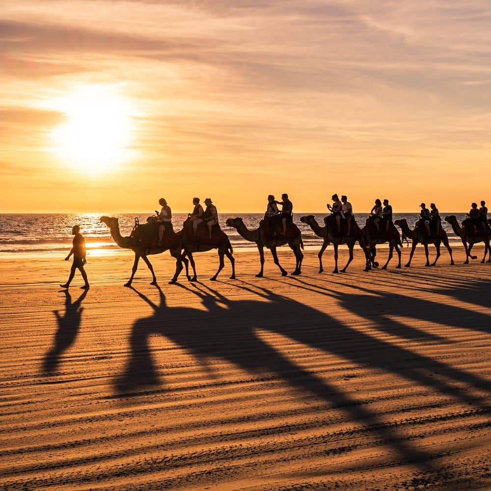 Silhouettes of a camel train and their riders cast long shadows on the sand of Cable Beach at sunset, with the golden sun dipping towards the horizon in Western Australia.