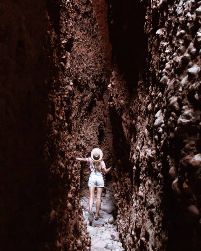 Beck of Salt and Charcoal in a sunhat and light clothing explores Echidna Chasm in Purnululu National Park, navigating through the narrow, towering walls of the deep red rock gorge.
