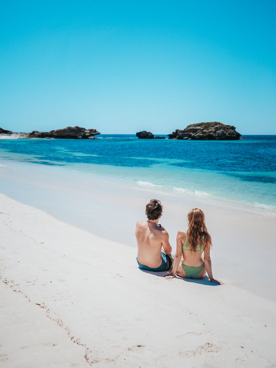 A couple sits closely together on the pristine white sands of a beach on Rottnest Island, Perth, looking out at the tranquil turquoise waters and rocky islets.