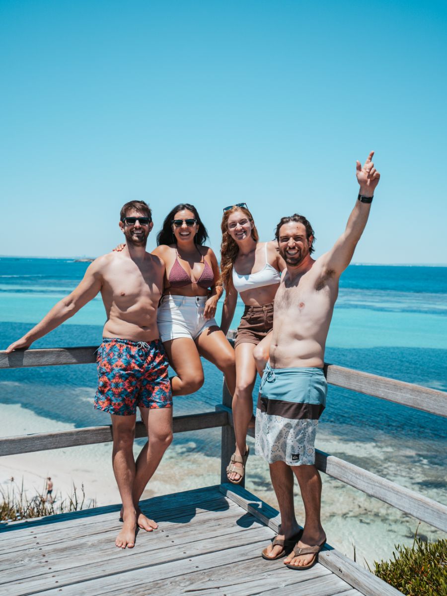 A group of friends in swimwear enjoys the sunny coast of Western Australia, posing on a wooden overlook against a backdrop of vibrant blue sea.