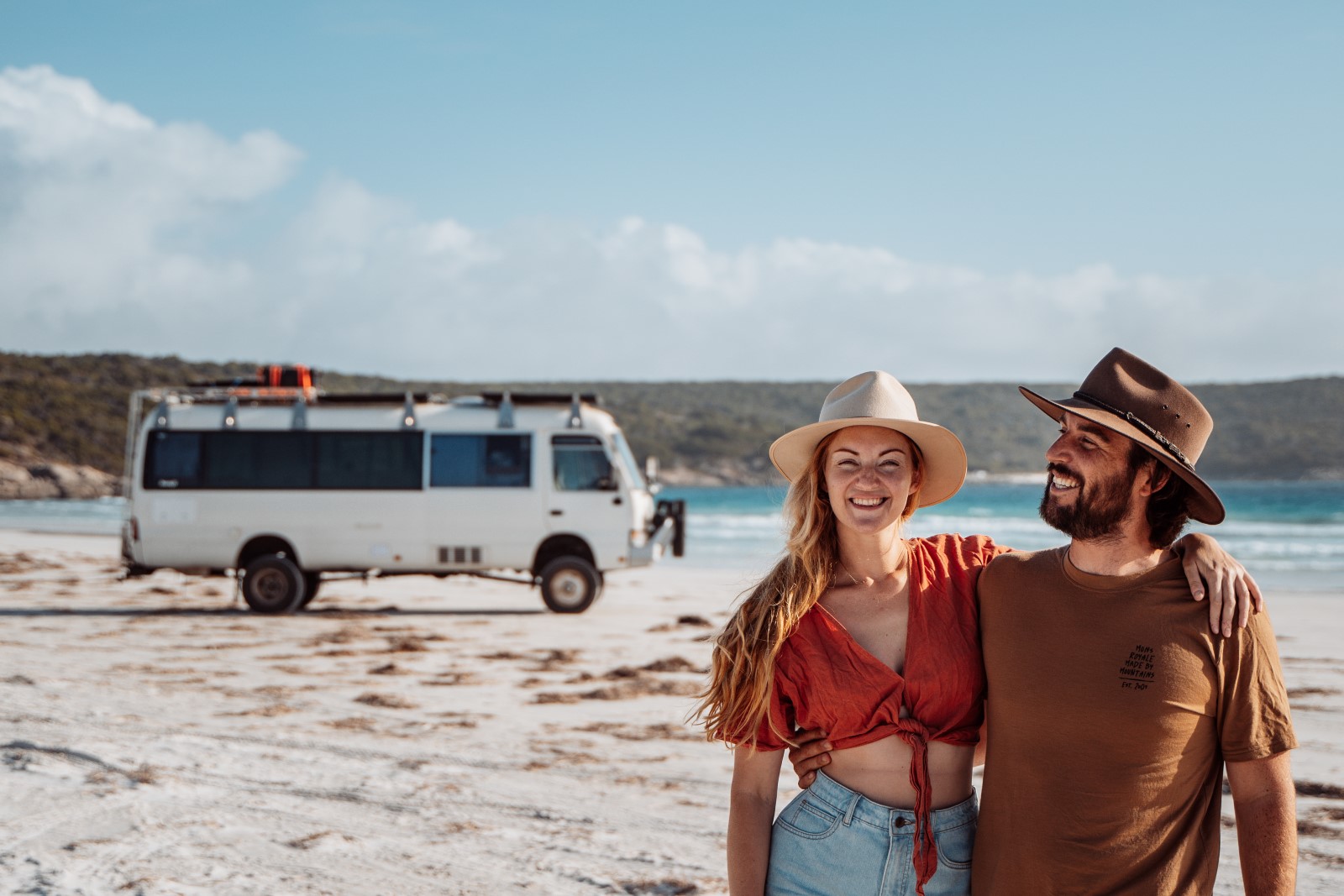 Alt text: A cheerful couple embraces in front of Salt and Charcoal  camper van parked on the sandy shore, with clear blue waters and a sunny sky in the background, evoking a sense of joy and adventure in Bremer Bay beach Western Australia