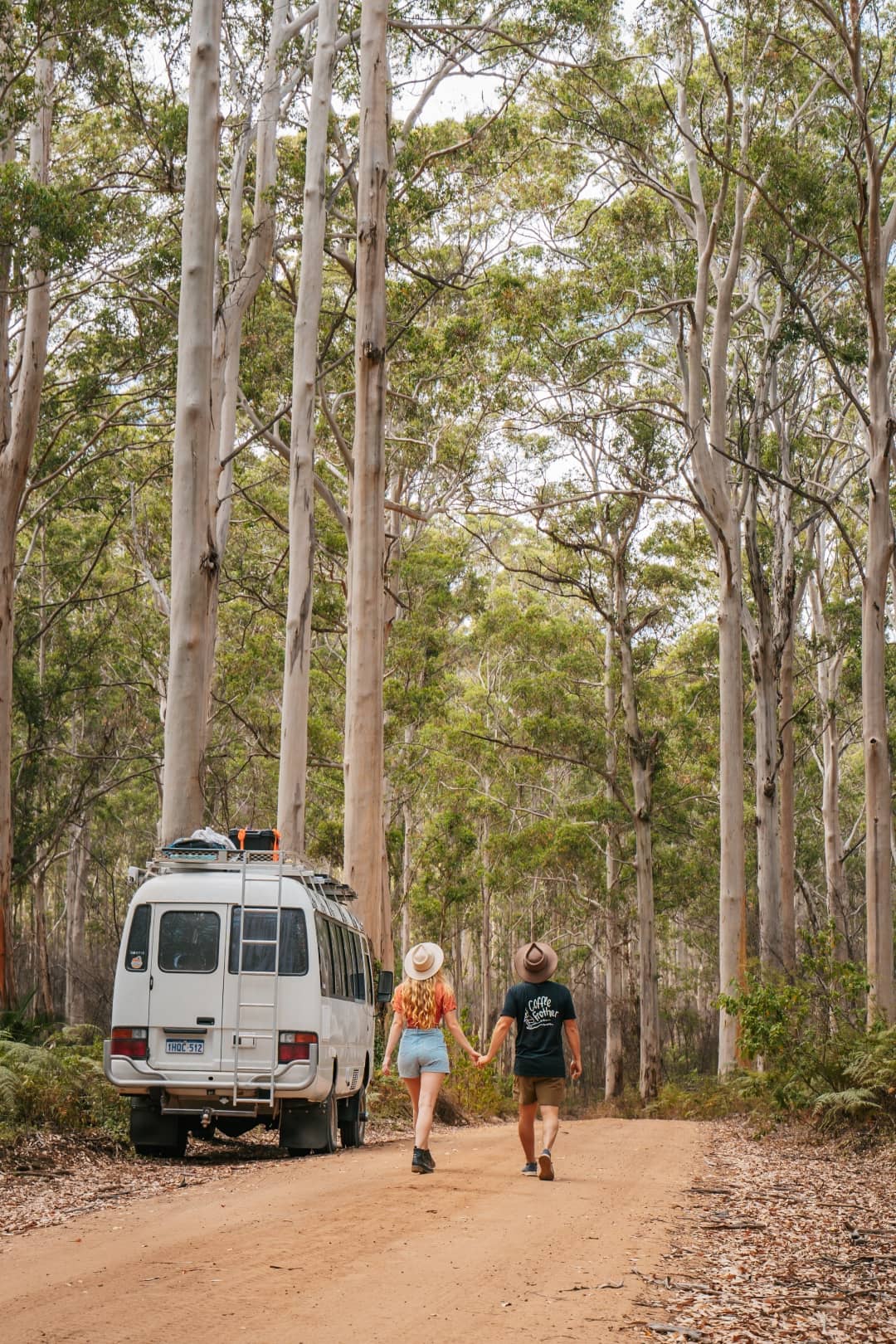 A couple beside a van holding hands while hiking in a forest with tall trees at the Boranup Forest, Margaret River, Western Australia
