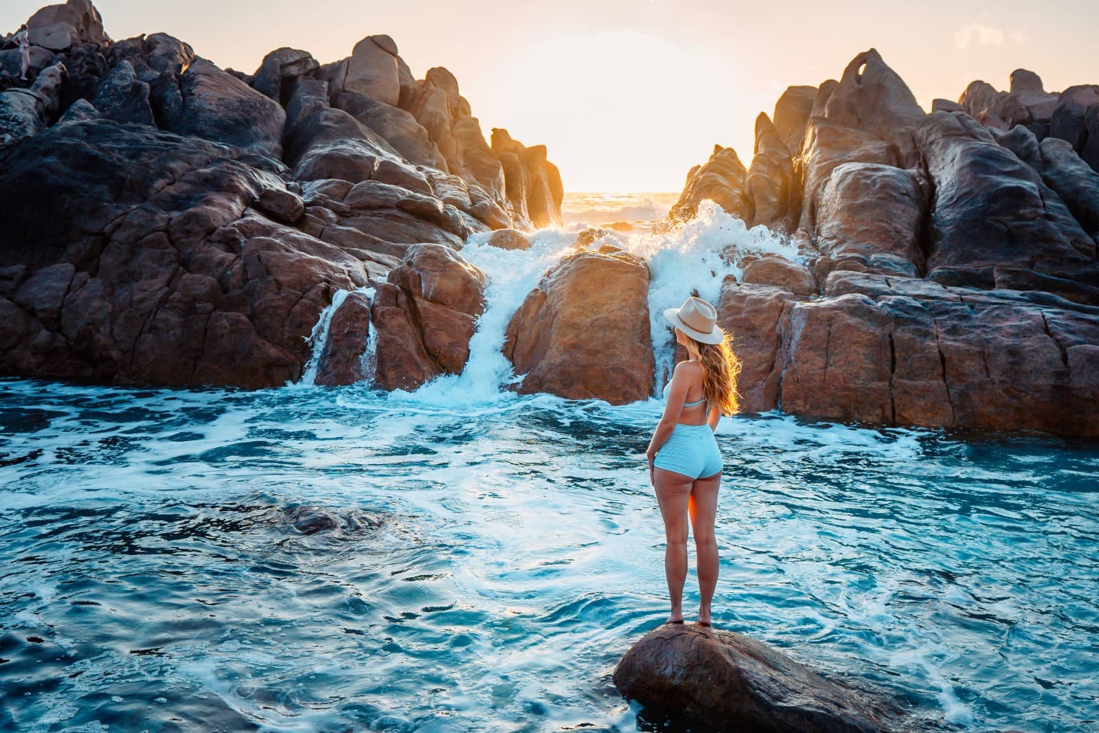 A person stands on a rock at the edge of Injidup Natural Spa in Yallingup, Western Australia, admiring the dynamic interaction of waves crashing against the rugged rocky shore at sunset