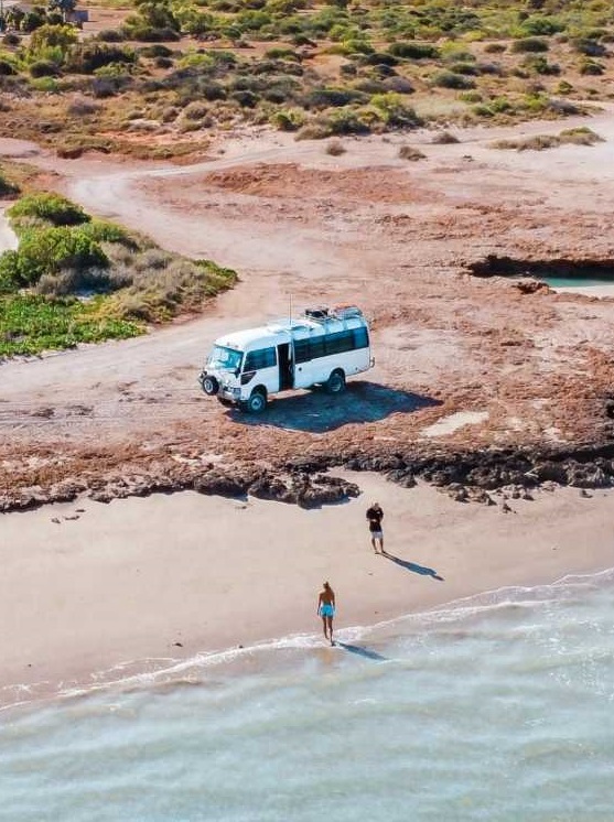 An aerial view of Salt and Charcoal camper van parked near the edge of a sandy beach in Western Australia, with two people enjoying the secluded shore by the crystal-clear water.