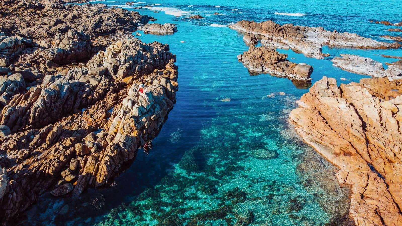 A bird's-eye view of the coastal formations at The Aquarium in Yallingup, Western Australia, showcasing clear, turquoise waters between rugged, orange-hued rocks, with a woman perched on the cliffs enjoying the view