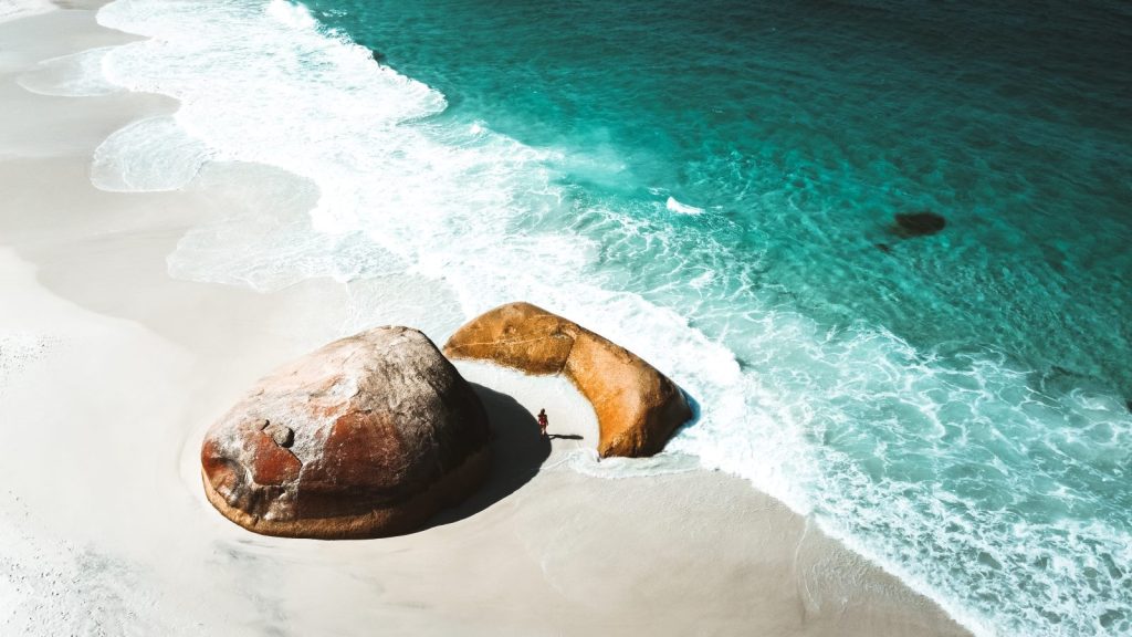 Aerial shot of a beach in Albany, Western Australia, with a person standing by large, reddish-brown rocks on white sand, bordered by turquoise waters.